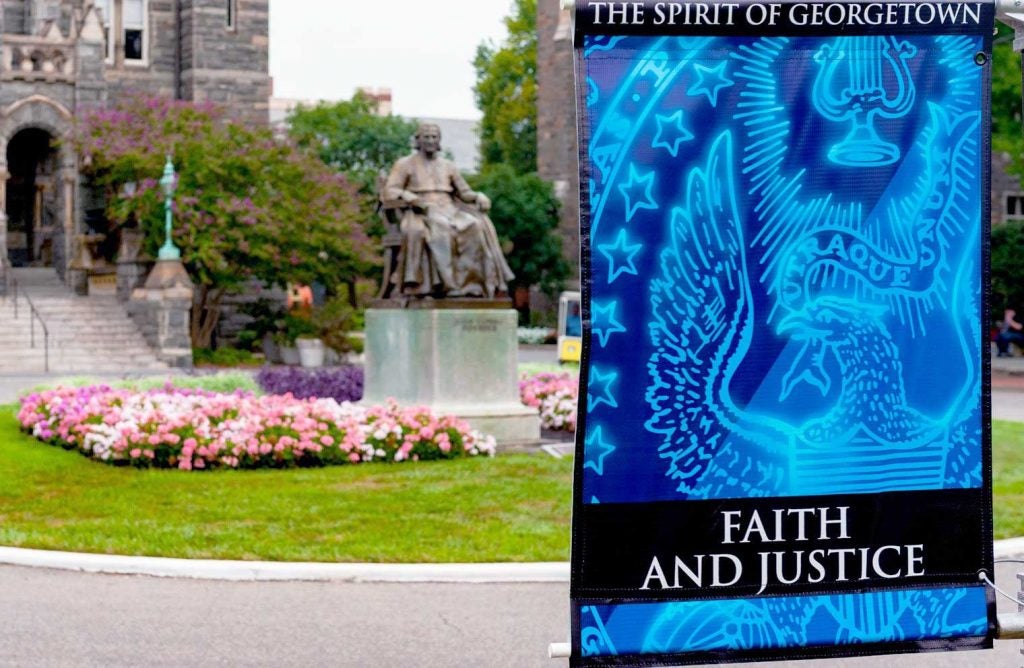 Image of John Carroll Statue, in front of Healy Hall, surrounded by flowering plants. In the foreground a banner with the words u0022Faith and Justiceu0022 and the GU Seal