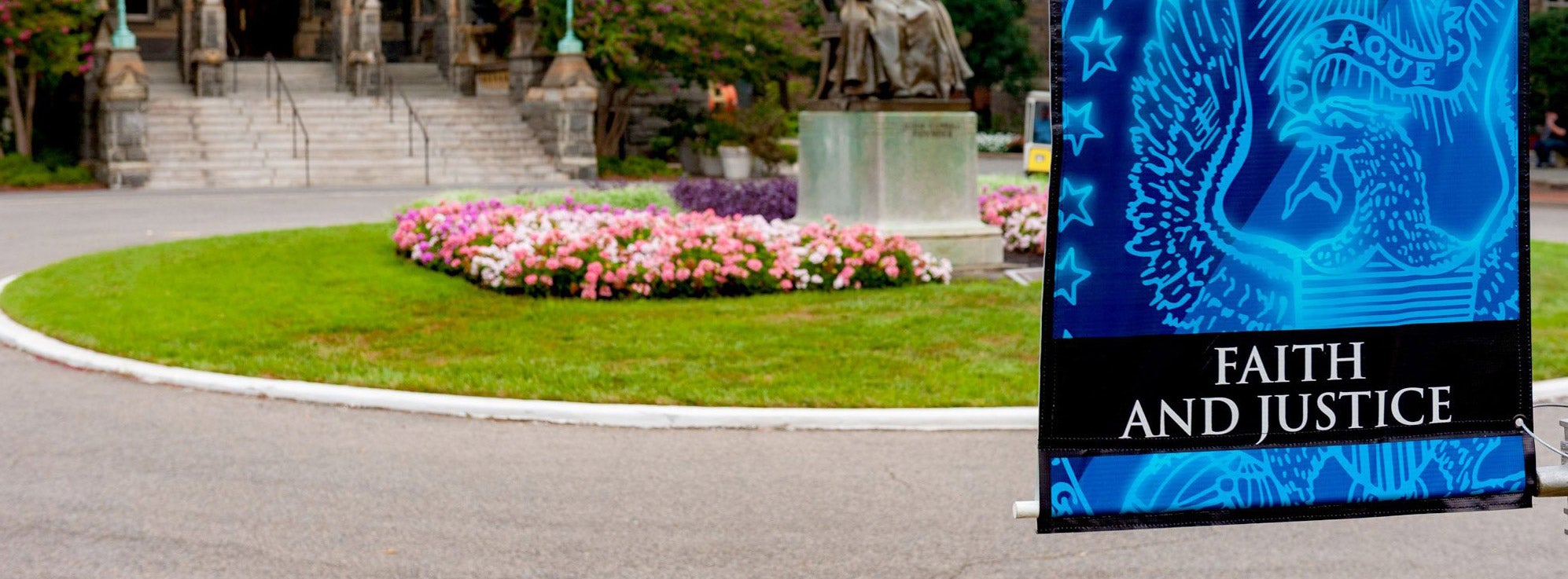 Image of John Carroll Statue, in front of Healy Hall, surrounded by flowering plants. In the foreground a banner with the words 